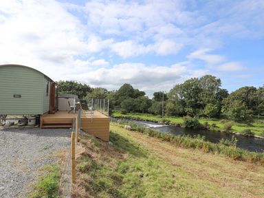 A green shepherds hut with a deck overlooking a river and grassy field with trees in the background at Llety'r Bugail 1 in Llanrug