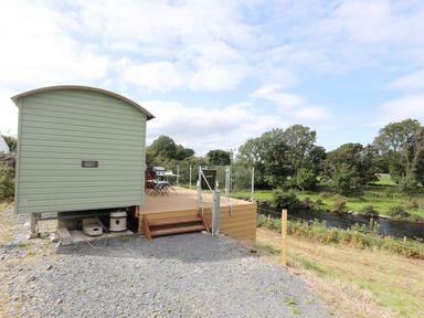 An outdoor area with a small green building wooden deck patio furniture and a river with trees in the background at Llety'r Bugail 1 in Llanrug