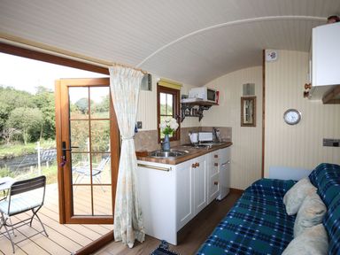 A compact kitchen with a sink microwave and toaster next to a blue patterned sofa and an open door leading outside at Llety'r Bugail 1 in Llanrug