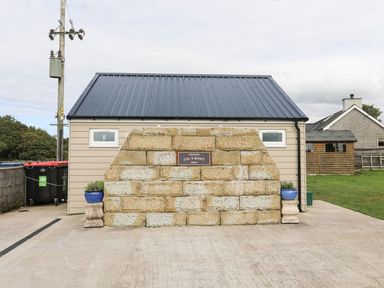 A stone wall with a plaque in front of a small building with a metal roof and two blue pots with plants at Llety'r Bugail 1 in Llanrug