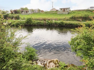 A river with bushes and rocks on the bank and houses on a hill at Llety'r Bugail 1 in Llanrug