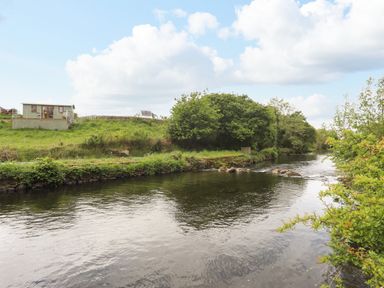 A river with trees and a grassy bank beside a small building and a vehicle at Llety'r Bugail 1 in Llanrug