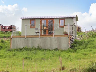 A small green cabin with glass railings and outdoor furniture on a raised platform with stairs surrounded by grass at Llety'r Bugail 1 in Llanrug