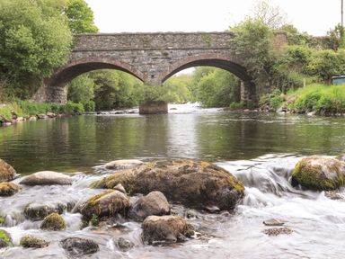 A stone arched bridge over a flowing river with rocks and trees at Llety'r Bugail 1 in Llanrug