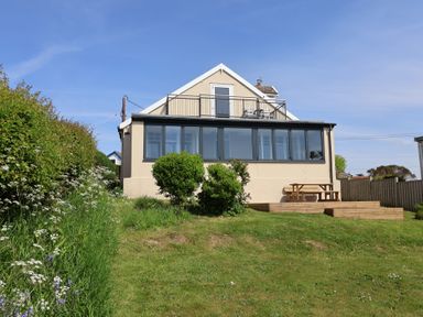 The back of a two-story house with a balcony and enclosed porch overlooking a grassy yard with bushes and a wooden picnic table on a deck at Tillicum in Charmouth