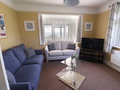 A living room with two sofas a glass coffee table a television on a stand and windows with curtains at Tillicum in Charmouth