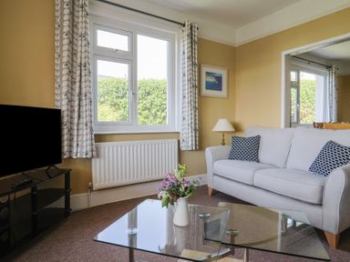 A living room with a white sofa and patterned cushions a glass coffee table with flowers a TV on a stand and curtains at Tillicum in Charmouth