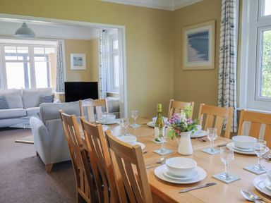 A dining room with a set dining table and an adjoining living room with sofas and a television at Tillicum in Charmouth