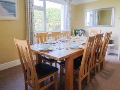 A dining room with a wooden table set for eight near a window with patterned curtains and a large mirror on the wall at Tillicum in Charmouth