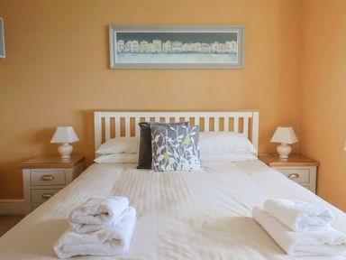 A bedroom with a double bed white bedding folded towels bedside tables with lamps and a framed painting above the bed at Tillicum in Charmouth