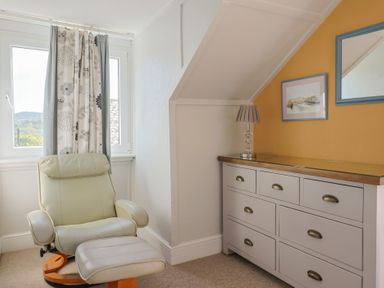 A room with a cream recliner chair by a window and a dresser with a lamp and framed pictures at Tillicum in Charmouth