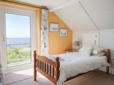 A bedroom with a wooden bed and white bedding next to a glass door overlooking the sea at Tillicum in Charmouth