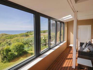 A sunroom with wicker sofas and small tables overlooking greenery and the sea at Tillicum in Charmouth