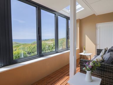 An enclosed porch with large windows overlooking greenery and the sea a wicker sofa with cushions a small table with a bottle and glasses and another table with a vase of flowers at Tillicum in Charmouth