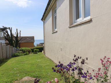 A side yard with grass and flowers next to a beige house with windows and a view of the sea at Tillicum in Charmouth
