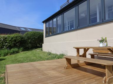 A wooden patio with a picnic table and benches next to a building with large windows and a garden with grass and bushes at Tillicum in Charmouth