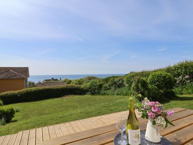 A garden with a wooden table holding a bottle of wine, two glasses, and a vase of flowers with a sea view at Tillicum in Charmouth
