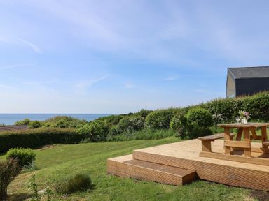 A wooden deck with picnic table and benches overlooking a grassy garden and sea at Tillicum in Charmouth