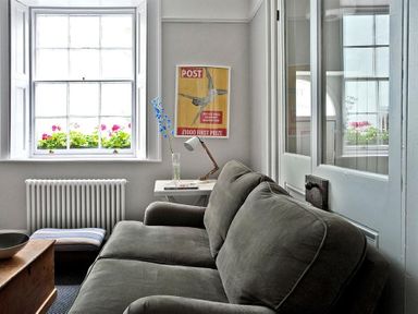 A sitting area with a gray sofa a side table with a lamp a window with flowers and a framed poster on the wall at The Arched House in Lyme Regis