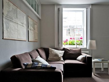 A living room with a brown sectional sofa and pillows near a window with flowers and a white side table with a lamp at The Arched House in Lyme Regis