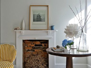 A room with a fireplace featuring exposed brick and a mantel with a framed picture and vases alongside a wooden round table with flowers and books and a yellow striped chair at The Arched House in Lyme Regis