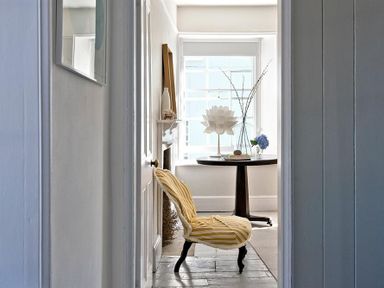 A hallway leading to a room with a yellow striped chair and a round table with decorative items by a window at The Arched House in Lyme Regis
