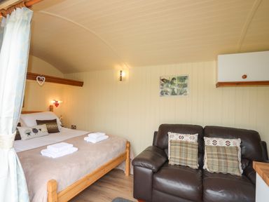 A bedroom with a wooden double bed with linens and folded towels next to a dark leather sofa with cushions at Llety'r Bugail 2 in Llanrug