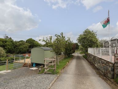 A narrow paved road with a Welsh flag on a pole next to a fenced stone wall and a green caravan beside a gravel path at Llety'r Bugail 2 in Llanrug