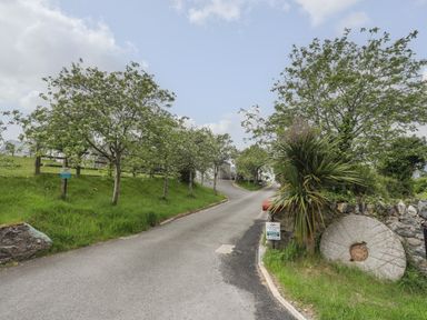 A rural road lined with trees and grass leading to buildings with a round stone and signs on the right side at Llety'r Bugail 2 in Llanrug