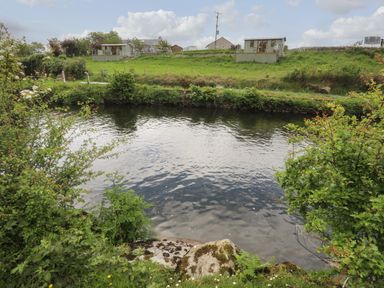 A river with grass and bushes on the banks and small houses in the background at Llety'r Bugail 2 Llanrug