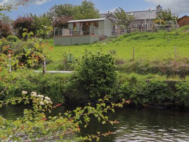 A green field with a small wooden structure and a house in the background near a river at Llety'r Bugail 2 in Llanrug