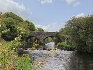 A stone bridge over a river with greenery and flowering plants at Llety'r Bugail 2 in Llanrug