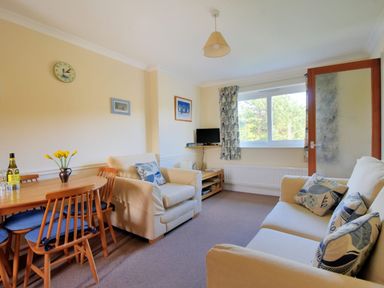 A living room with a wooden dining table and chairs two beige sofas with patterned cushions a small TV on a stand and a window with patterned curtains at Bridge House Apartment in Charmouth