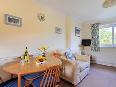 A dining table with a bottle of wine and fruit bowl next to an armchair and a small TV under a window at Bridge House Apartment in Charmouth