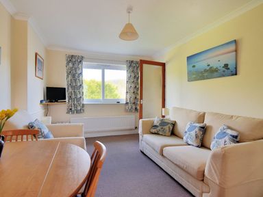 A living room with a beige sofa and armchair with patterned cushions a round wooden dining table chairs a small TV on a wall shelf and a window with curtains at Bridge House Apartment in Charmouth