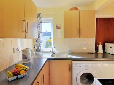 A kitchen with wooden cabinets a kettle a bowl of fruit a washing machine and a window at Bridge House Apartment in Charmouth