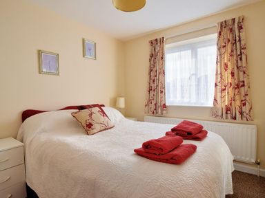 A bedroom with a bed with white bedding and red towels on top next to a window with patterned curtains at Bridge House Apartment in Charmouth
