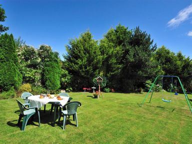 A garden with plastic chairs and table set on grass surrounded by trees and a swing set in Bridge House Apartment in Charmouth