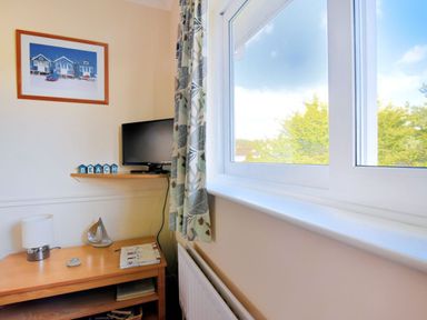 A corner of a room with a window and curtains a wooden table with a lamp a silver sailboat figurine books a wall-mounted tv and a framed picture at Bridge House Apartment in Charmouth