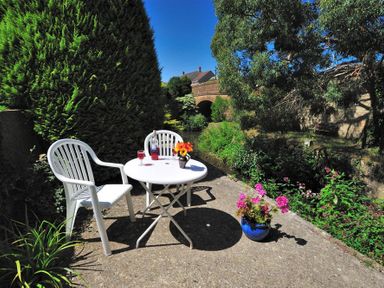 A small outdoor patio with a white table and two white chairs near a garden with flowers and trees Bridge House Apartment Charmouth