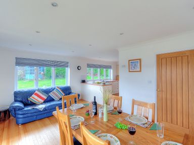 A dining area with a wooden table set with plates and glasses next to a blue sofa in an open living room and kitchen at Pendower in Charmouth