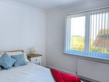 A bedroom with a bed with blue pillows a bedside table with a lamp a window with vertical blinds and a radiator at Pendower in Charmouth