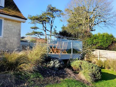A garden with shrubs and grass surrounding a glass balcony with wooden chairs at Pendower in Charmouth