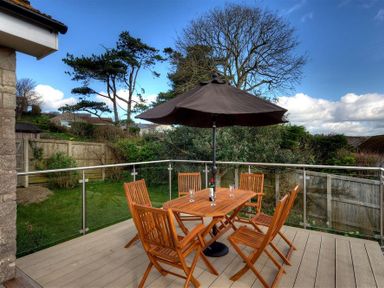 A wooden patio table with six chairs and a black umbrella on a deck overlooking a garden at Pendower in Charmouth