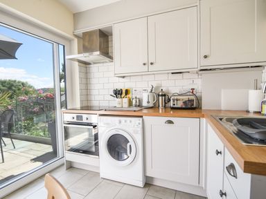 A kitchen with white cabinets wooden countertops a washing machine oven toaster kettle and kitchen utensils next to a glass door with a view of a garden at Nutwood in Charmouth