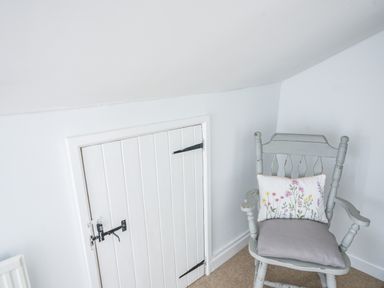 A small white wooden door with black latch beside a wooden chair with a floral cushion in a white room at Nutwood in Charmouth