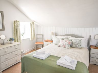 A bedroom with a bed with floral pillows a green blanket a chair two bedside tables lamps a dresser with a mirror and flowers at Nutwood in Charmouth