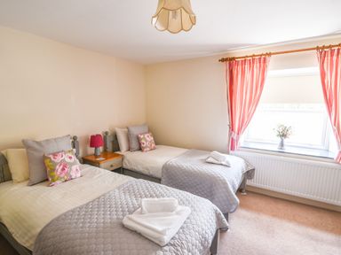 A bedroom with two single beds with gray quilts and floral pillows and a window with red-striped curtains at Nutwood in Charmouth