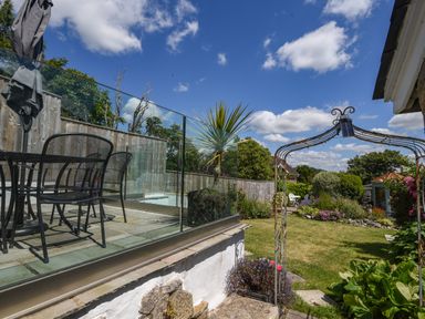 A garden with a metal table and chairs on a patio behind a glass fence and a metal archway leading to a grassy area with plants and flowers at Nutwood in Charmouth