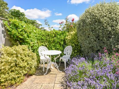 A small outdoor patio area with a white metal table and two chairs surrounded by greenery and flowers at Nutwood in Charmouth